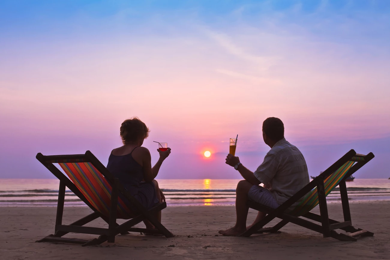 A couple sitting on deckchairs on the beach with cocktails in their hands watching the sunset over the ocean