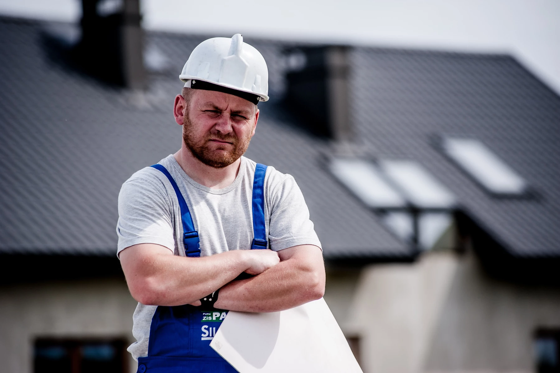 A man wearing a hardhat and work gear standing in-front of a building with a rolled up paper sheet with his arms crossed