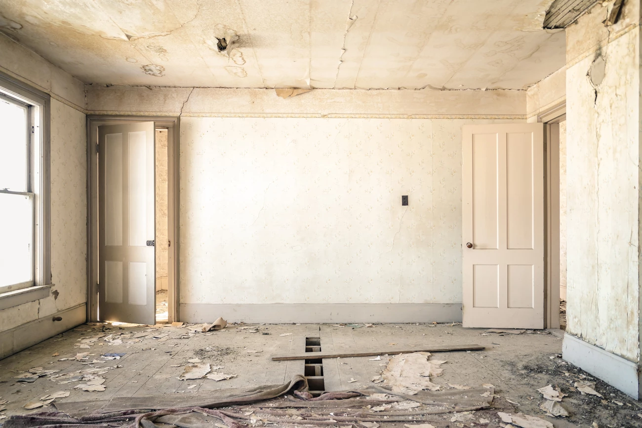 A dilapidated front room of a house with broken floor boards and peeling wallpaper 