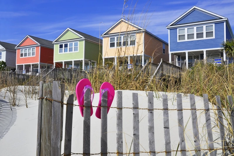 Pink flip flops hung on a thin wooden fence on a sandy beach in-front of four different coloured houses