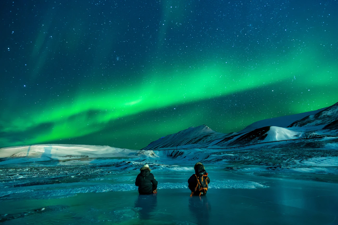 Two people sitting on the ground watching the northern lights over a snowy region