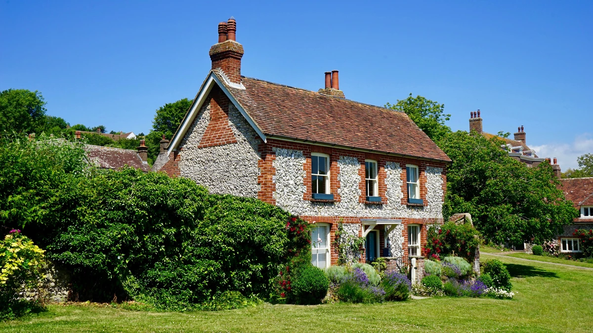 A stone holiday cottage in a green garden surrounded by bushes and flowers on a sunny day