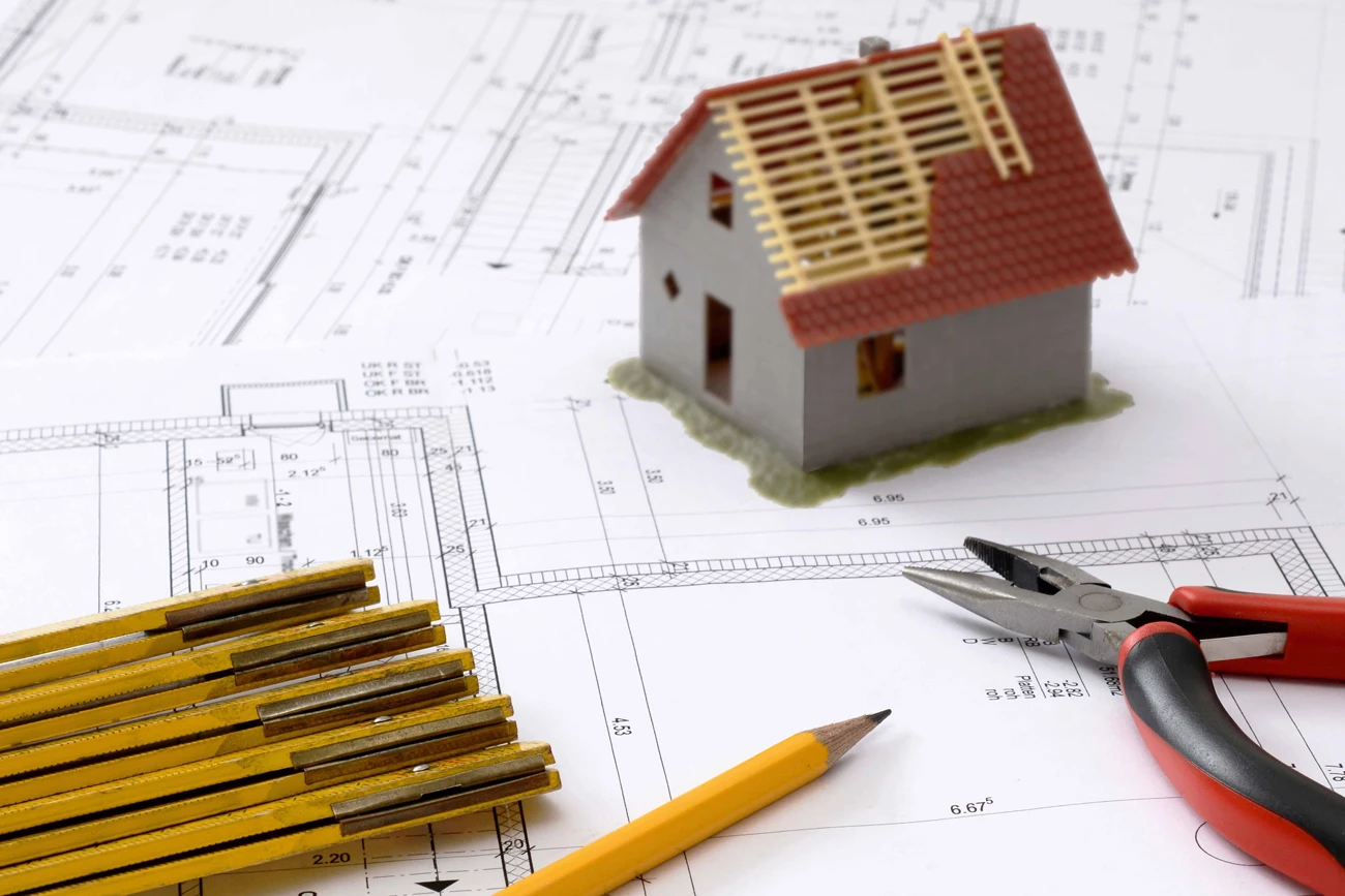 A desk with building plans laid across it as well as pencils and a model of a house under construction