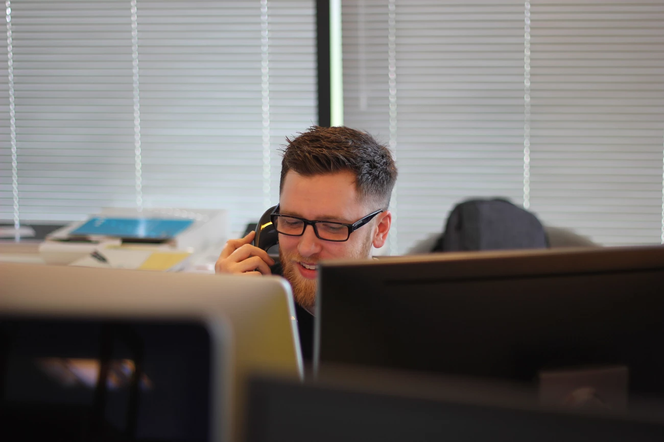 A man in an office talking on a phone whilst typing into a computer