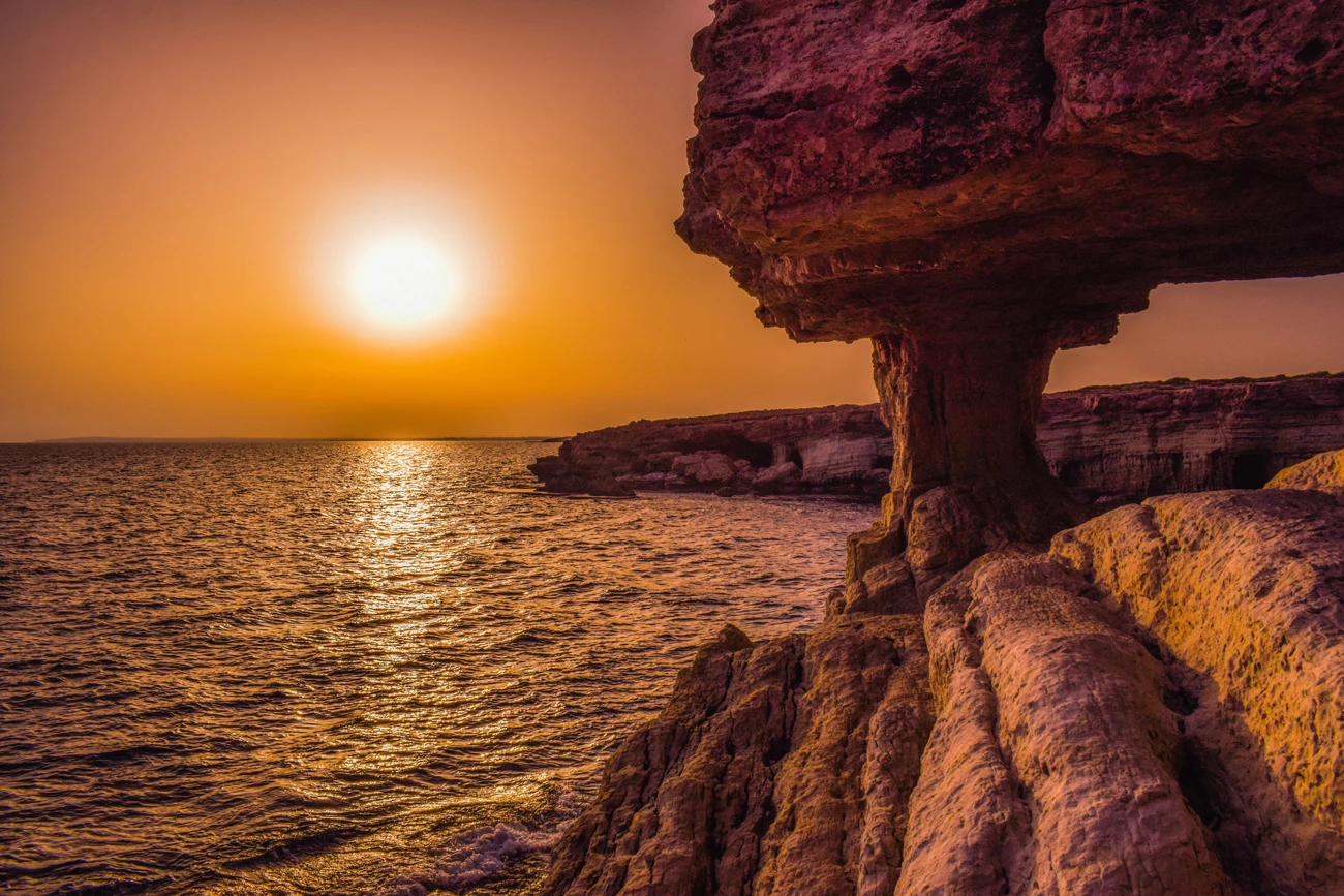 The bright orange sun sets over the ocean around Cyprus with the rocky shoreline and a rock arch in the foreground
