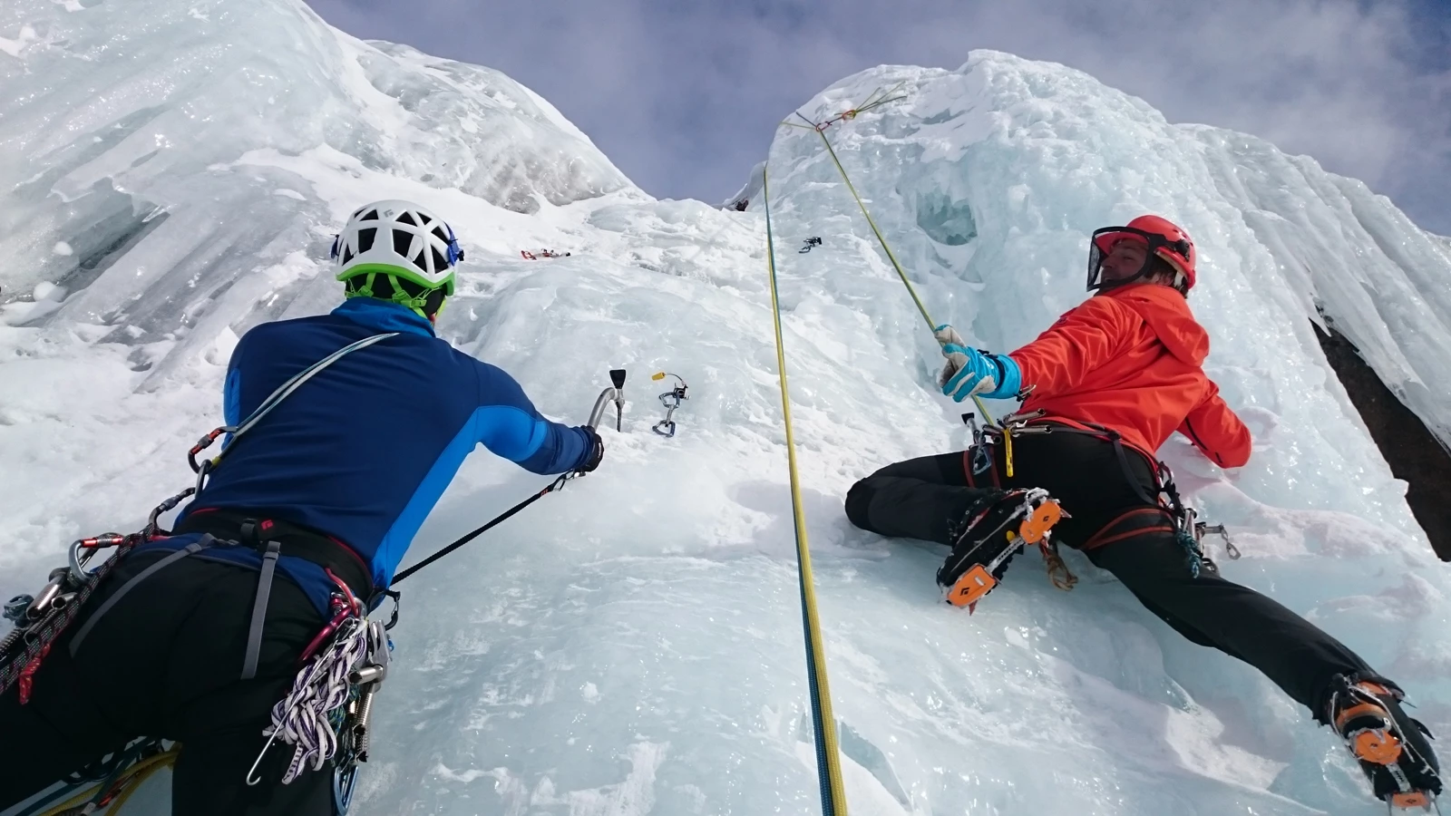 Two people in winter clothing climbing a wall of ice using tools and ropes