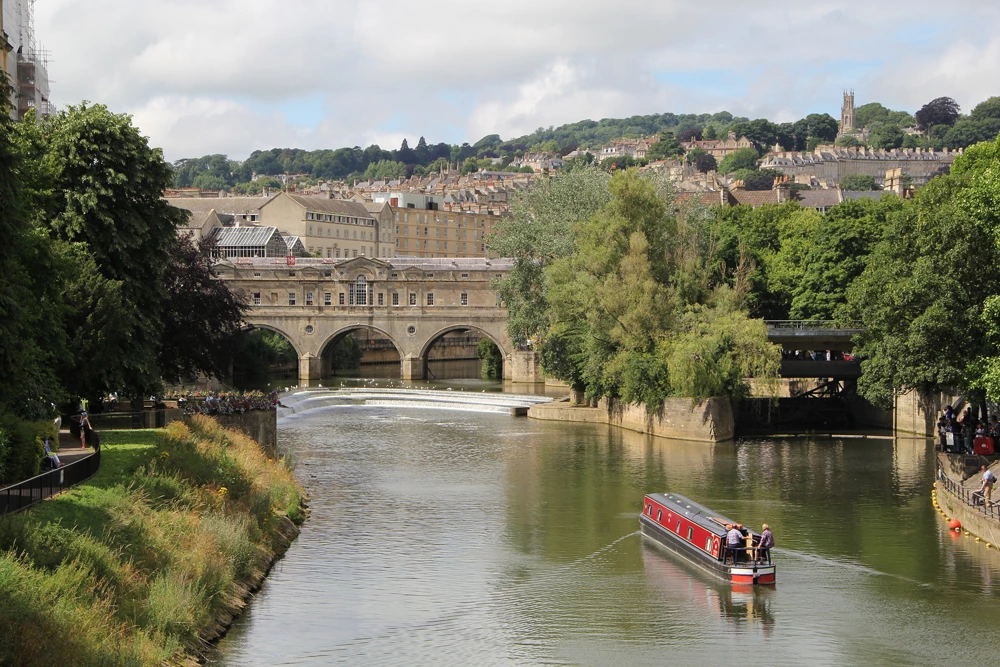 A view over Bath with a stunning bridge and wide river running under with a small canal boat heading towards it