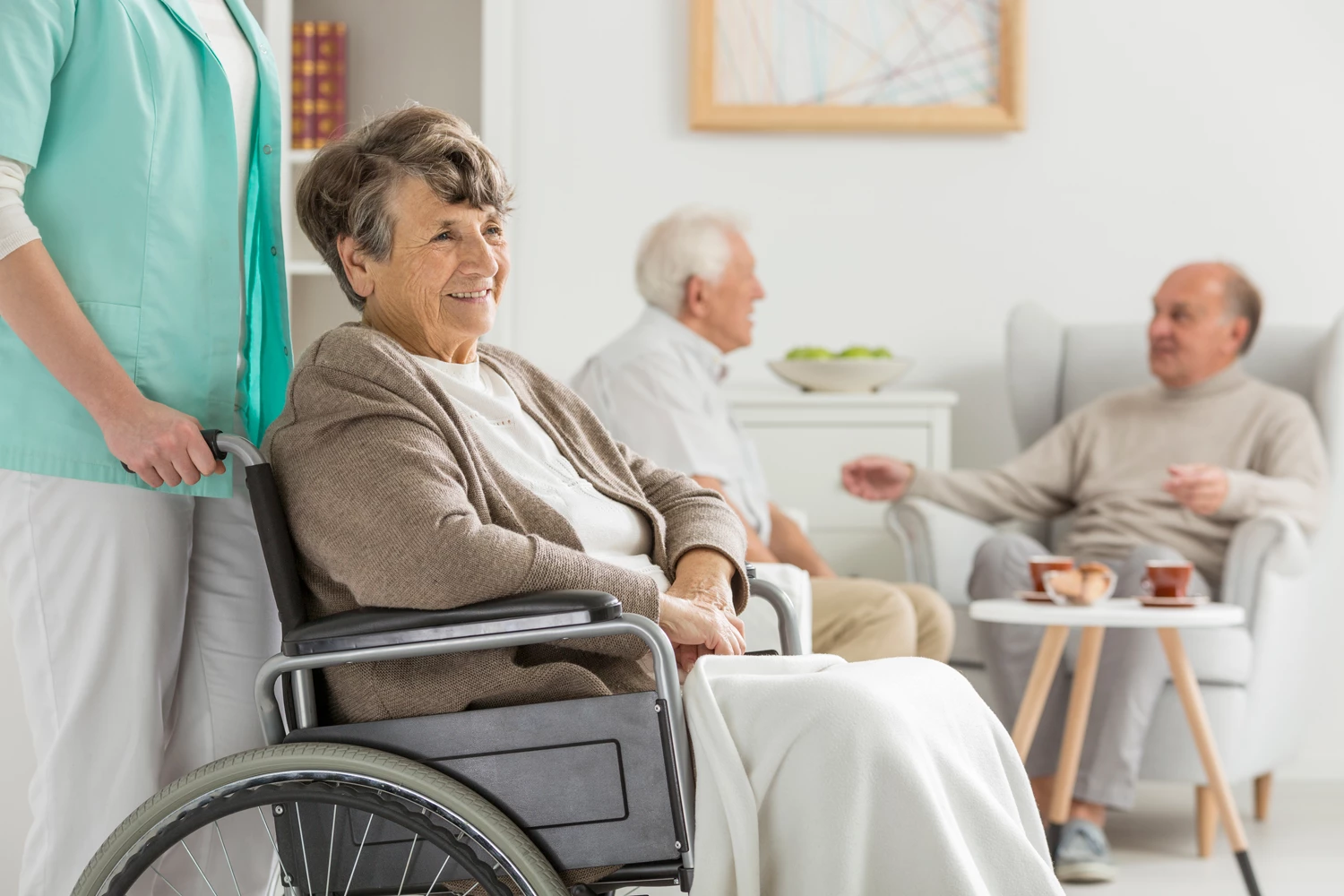 A care worker pushing an elderly lady in a wheelchair through a care home lounge area