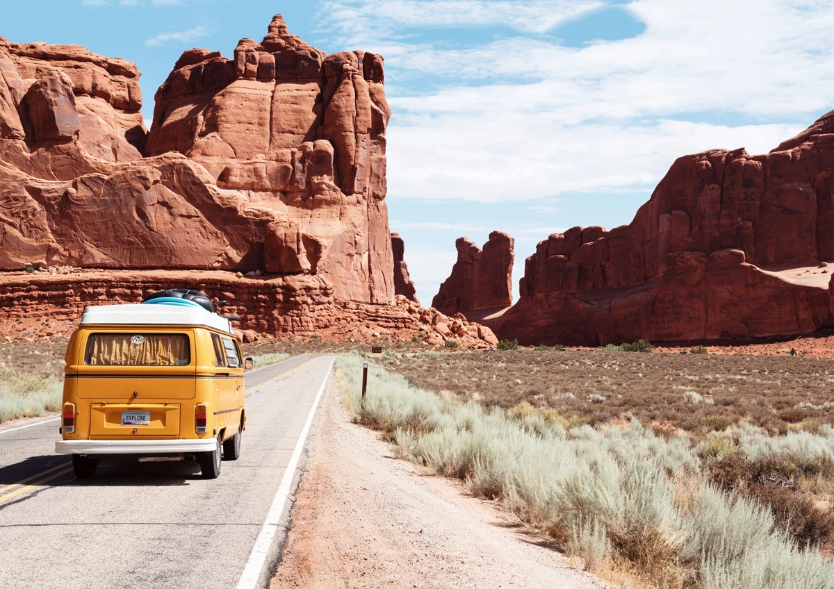 A yellow campervan driving on an open road through red cliffs in the USA