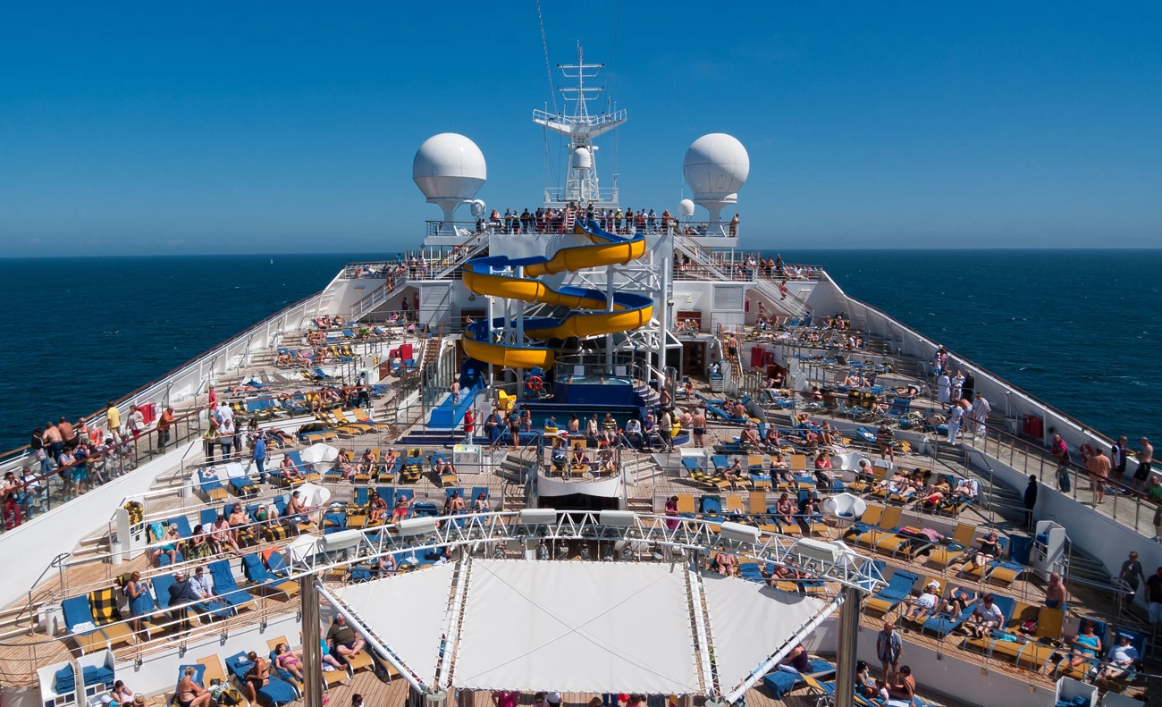 The crowded deck at the rear of a cruise ship with two water slides at the end